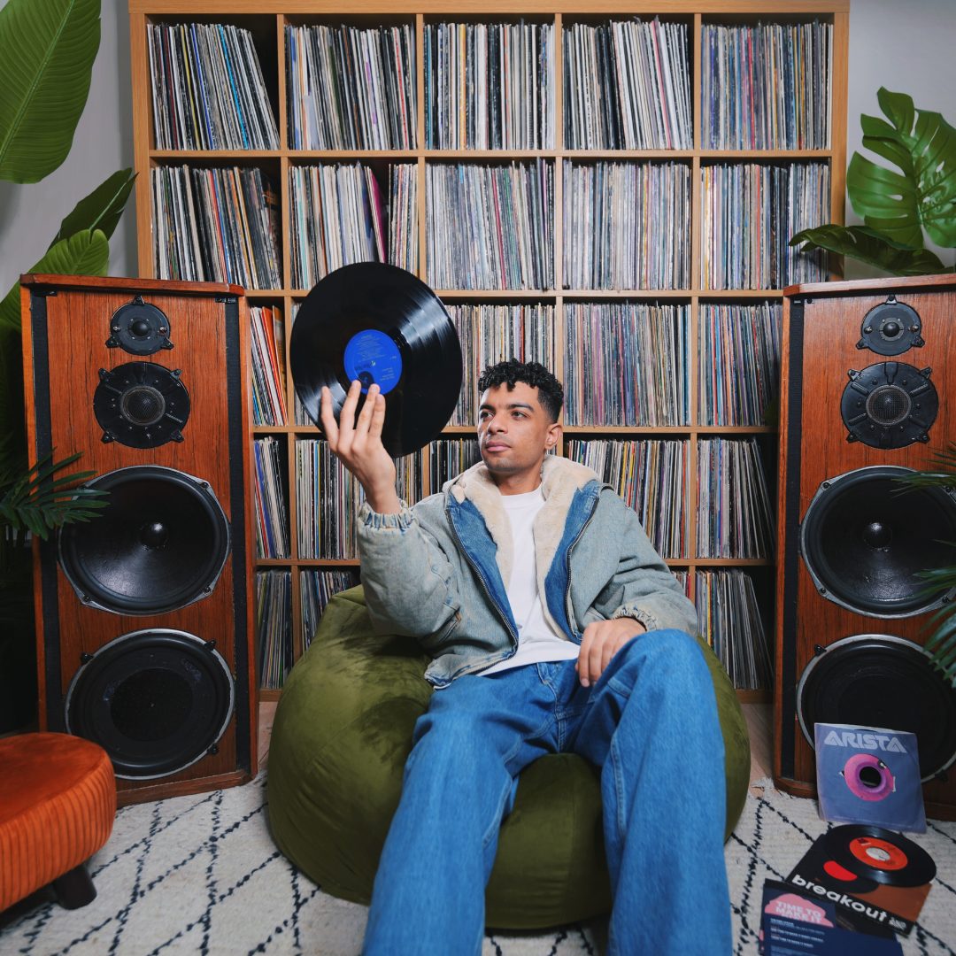 Kirollus on a green beanbag holding up a record, in front of shelves full of vinyl and two wooden speakers