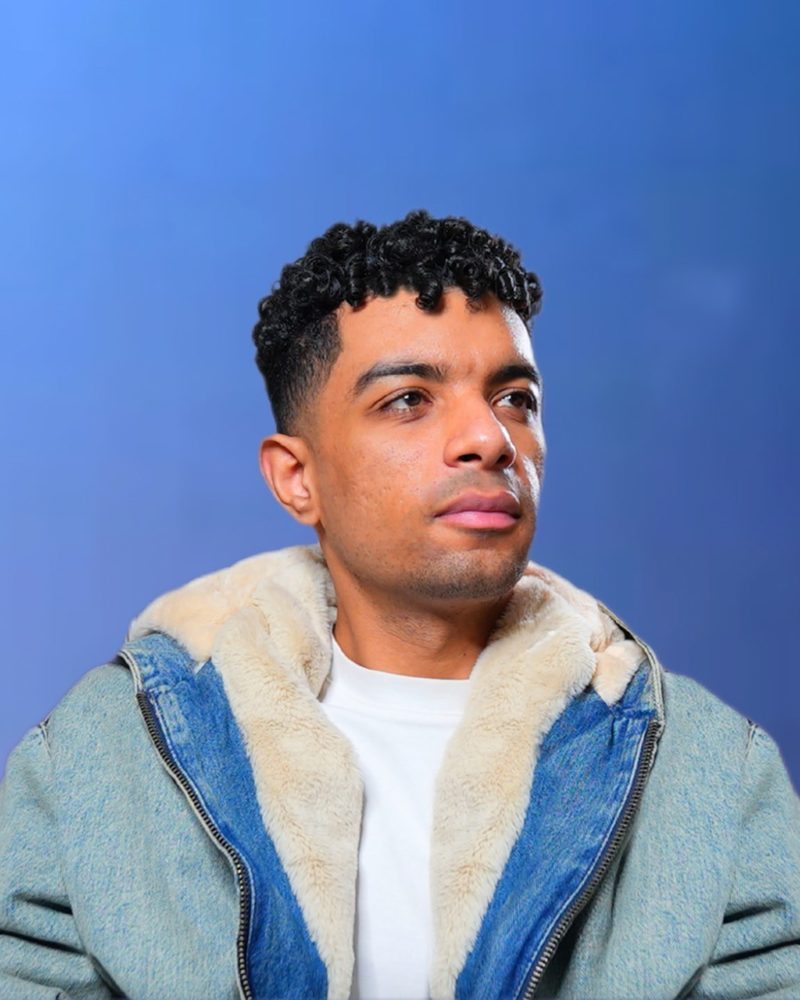Portrait of Kirollus in a fleece-lined denim jacket against a blue backdrop