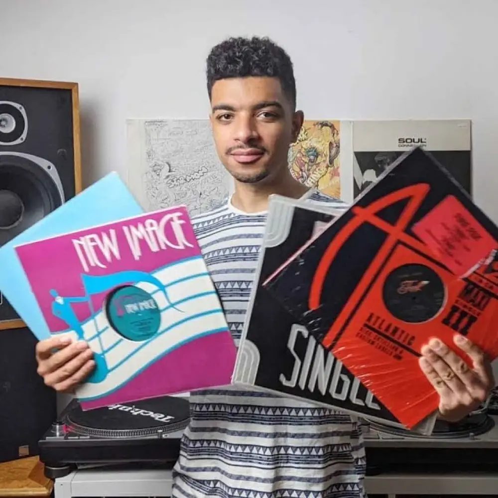 Kirollus holding up four 12-inch sleeves, including New Image and an Atlantic maxi single, in front of his turntables