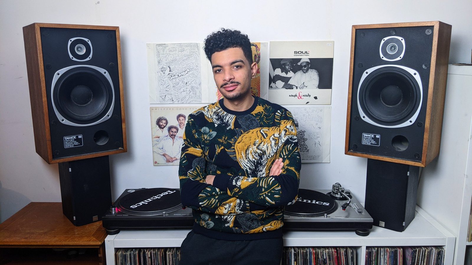Kirollus with arms crossed in front of two Technics turntables, Tannoy Oxford speakers and record sleeves pinned to the wall